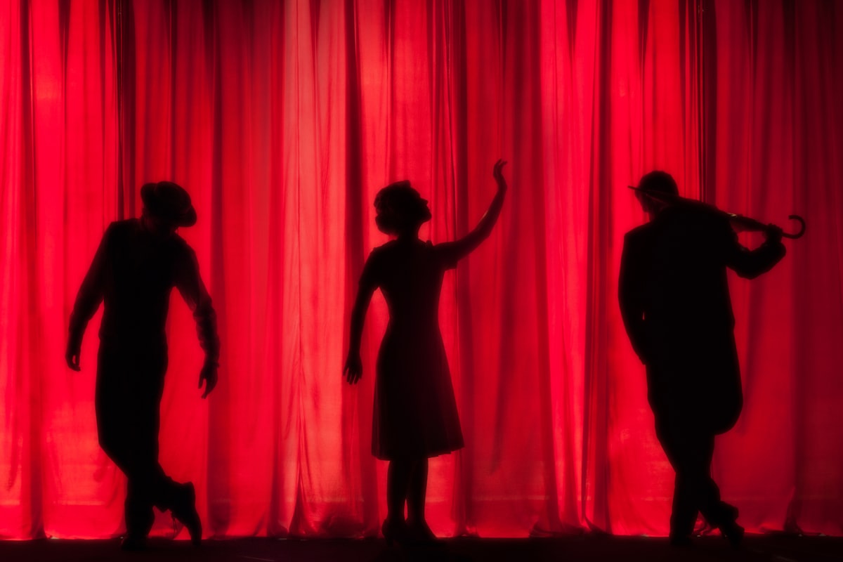 Schauspielerinnen und Schauspieler als Silhouetten vor rotem Bühnenvorhang.